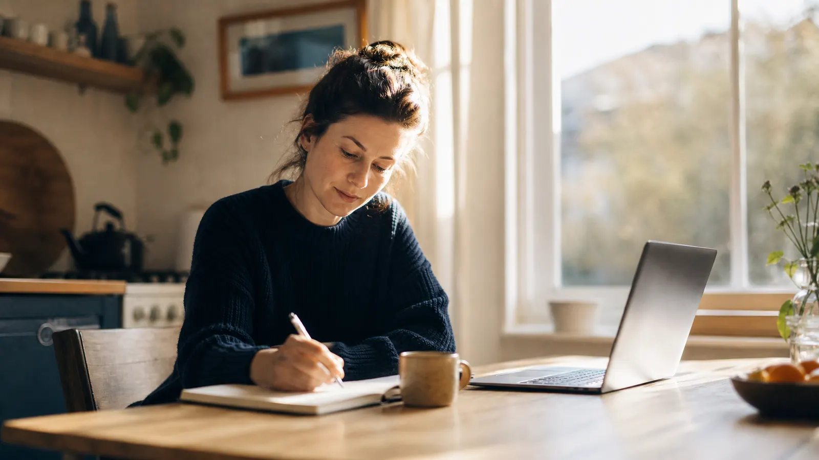 A woman writing notes at a sunlit kitchen table with a laptop open beside her